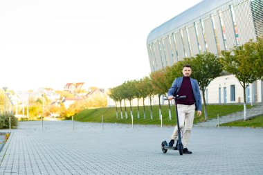 Gün batımında havaalanının yanındaki kaldırımda e-scooterlı genç yakışıklı adam. Modern elektrikli scooter 'la son moda şehir ulaşımı. Çevre dostu hareketlilik kavramı