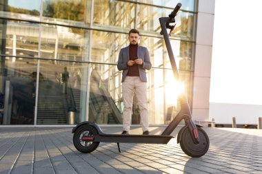 Günbatımında havaalanının yanındaki kaldırımda duran ve akıllı telefondan konuşan, e-scooter 'lı genç yakışıklı bir adam. Modern elektrikli scooterla trend şehir ulaşımı. Çevre dostu hareketlilik konsepti.