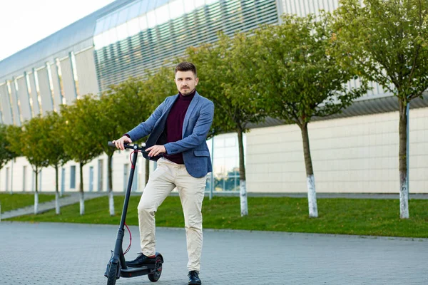 Young handsome stylish man with e-scooter standing on sidewalk near the airport on the sunset. Trendy urban transportation on modern electric scooter. Eco friendly mobility concept — Stock Photo, Image
