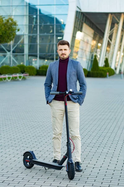 Young handsome stylish man with e-scooter standing on sidewalk near the business center on the sunset. Trendy urban transportation on modern electric scooter. Eco friendly mobility concept — Stock Photo, Image