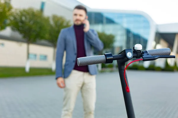 Junger gutaussehender Mann mit E-Scooter, der bei Sonnenuntergang auf dem Bürgersteig in der Nähe des Flughafens steht und mit dem Smartphone spricht, trendiger Stadtverkehr auf modernem Elektroroller. Umweltfreundliches Mobilitätskonzept — Stockfoto