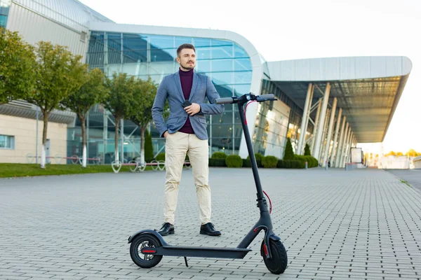 Ung stilig stilig man med e-scooter står på trottoaren nära flygplatsen vid solnedgången. Trendig stadstransport på modern elskoter. Miljövänlig rörlighet — Stockfoto