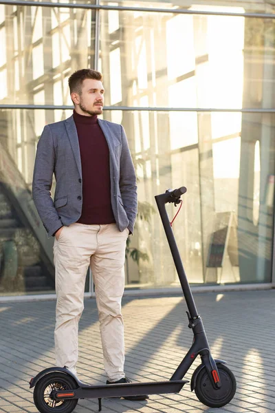 Młody, przystojny, stylowy mężczyzna z e-skuterem stojący na chodniku w pobliżu centrum biznesowego przy zachodzie słońca. Modny transport miejski na nowoczesnym skuterze elektrycznym. Koncepcja mobilności ekologicznej — Zdjęcie stockowe