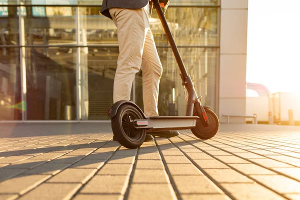 Egy férfi lábai állnak az e-robogón, ami a járdán parkol a városkép előtt a naplementében. Trendi városi közlekedés modern elektromos robogóval. Környezetbarát mobilitás koncepciója — Stock Fotó