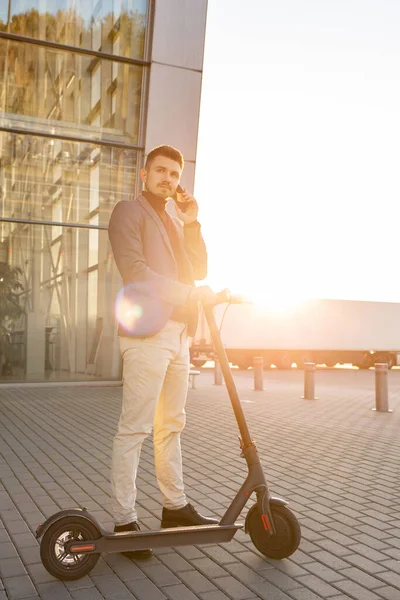 Junger gutaussehender Mann mit E-Scooter steht bei Sonnenuntergang auf dem Bürgersteig in der Nähe des Flughafens und telefoniert mit dem Smartphone. Trendiger Stadtverkehr auf modernem Elektroroller, umweltfreundliches Mobilitätskonzept — Stockfoto