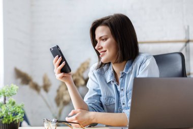 Evden çalışıyorum. Meslektaşlar ve serbest çalışanlarla çevrimiçi iletişim. Akıllı telefon kullanan ve gülümseyen genç bir kadının portresi. Cep telefonu uygulamaları, mesajlar, internet tarayıcıları, akıllı telefon aramaları.