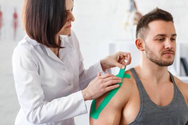 Kineziyoloji bandı. Fizyoterapist hastanın omzuna kinezyoloji bandı uyguluyor. Erkek sporcunun yaralı omzunu tedavi eden kadın terapist. Travma sonrası rehabilitasyon, spor fizyoterapisi