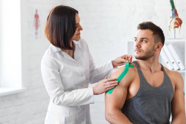 Kineziyoloji bandı. Fizyoterapist hastanın omzuna kinezyoloji bandı uyguluyor. Erkek sporcunun yaralı omzunu tedavi eden kadın terapist. Travma sonrası rehabilitasyon, spor fizyoterapisi