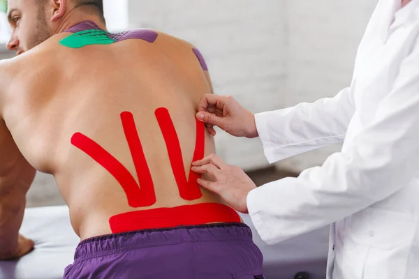 Kinesiologie-Aufnahmen. Physiotherapeut, der Kinesiologie-Klebeband auf den Rücken des Patienten aufträgt. Therapeut behandelt verletzten unteren Rücken eines jungen männlichen Athleten. Posttraumatische Rehabilitation, Sportphysikalische Therapie — Stockfoto