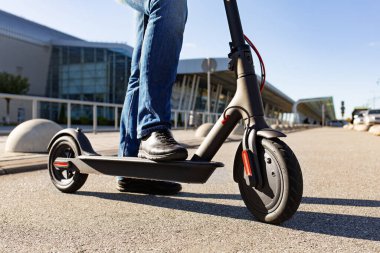 Gün batımında şehir manzarasında kaldırıma park etmiş, e-scooter 'ın üzerinde duran bir adamın bacakları. Modern elektrikli scooter 'la son moda şehir ulaşımı. Çevre dostu hareketlilik kavramı