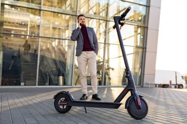 Günbatımında havaalanının yanındaki kaldırımda duran ve akıllı telefondan konuşan, e-scooter 'lı genç yakışıklı bir adam. Modern elektrikli scooterla trend şehir ulaşımı. Çevre dostu hareketlilik konsepti.
