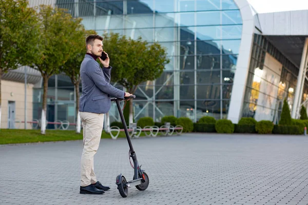 Günbatımında havaalanının yanındaki kaldırımda duran ve modern elektrikli scooterla trend şehir ulaşımı hakkında konuşan, e-scooter 'lı genç yakışıklı bir adam. Çevre dostu hareketlilik kavramı. — Stok fotoğraf