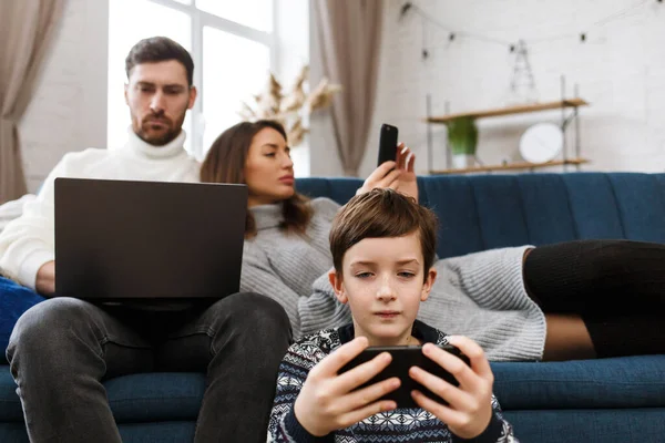 Mother, father and son using laptop and mobile phones at home. Family members ignoring each other and live talk. Gadget influence on family relationships. Modern technologies and addiction concept