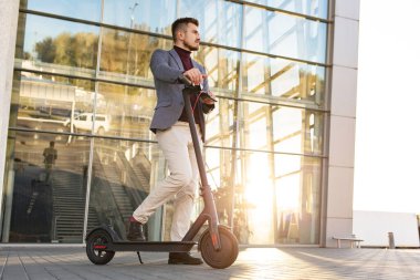 Günbatımında iş merkezinin yanındaki kaldırımda e-scooterlı genç yakışıklı adam. Modern elektrikli scooter 'la son moda şehir ulaşımı. Çevre dostu hareketlilik kavramı