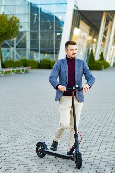 Günbatımında iş merkezinin yanındaki kaldırımda e-scooterlı genç yakışıklı adam. Modern elektrikli scooter 'la son moda şehir ulaşımı. Çevre dostu hareketlilik kavramı — Stok fotoğraf