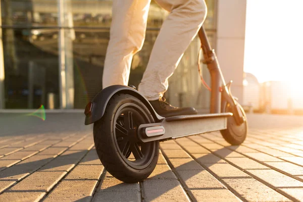 Die Beine eines Mannes, der auf einem E-Roller steht, der auf dem Bürgersteig am Stadtbild bei Sonnenuntergang abgestellt ist. trendige Stadtverkehr auf modernen Elektroroller. Umweltfreundliches Mobilitätskonzept — Stockfoto