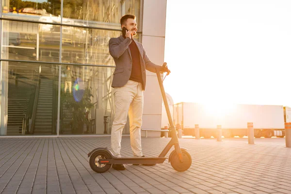 Joven hombre guapo con e-scooter de pie en la acera cerca del aeropuerto en la puesta del sol y hablando en el smartphone.Trendy transporte urbano en scooter eléctrico moderno, concepto de movilidad ecológica —  Fotos de Stock