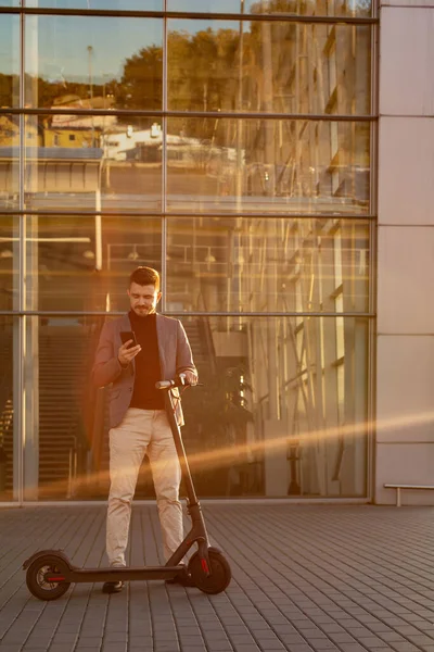 Young handsome man with e-scooter standing on sidewalk near the airport on the sunset and talking on the smartphone.Trendy urban transportation on modern electric scooter,Eco friendly mobility concept — Stock Photo, Image