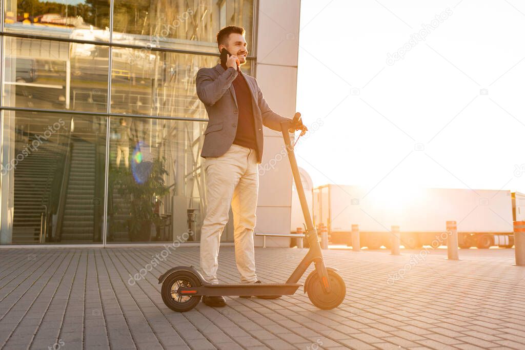 Young handsome man with e-scooter standing on sidewalk near the airport on the sunset and talking on the smartphone.Trendy urban transportation on modern electric scooter,Eco friendly mobility concept
