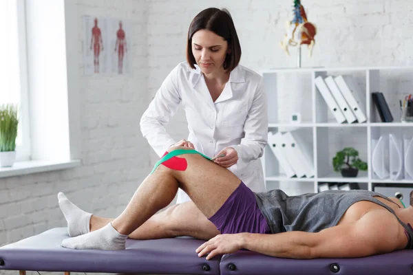 Fizjoterapeuta stosujący taśmę kinezjologiczną do kolana pacjenta.Terapeuta leczący uraz kolana młodego sportowca.Rehabilitacja pourazowa, rehabilitacja sportowa, koncepcja rekonwalescencji — Zdjęcie stockowe