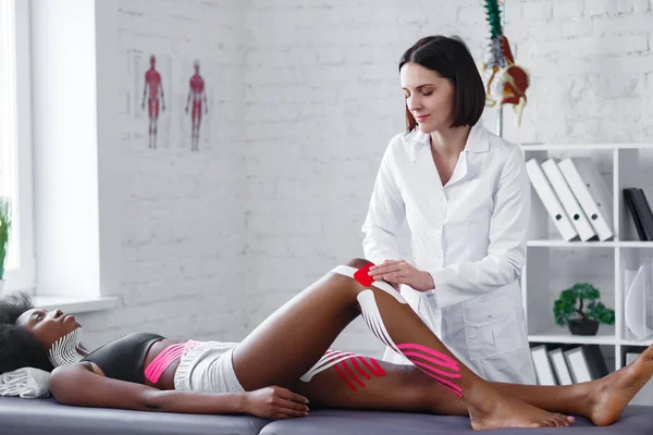 Fyzioterapeut aplikuje kineziologickou pásku na kolena pacienta.Terapeut léčí mladou afroamerickou atletku. Kineziologické nahrávání. Posttraumatická rehabilitace, sportovní rehabilitace — Stock fotografie