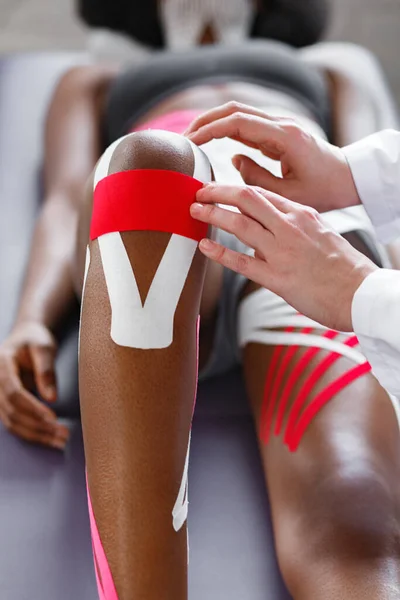 Sjukgymnast tillämpar kinesiologi tejp på patientens knä. Terapeut behandla unga kvinnliga afroamerikanska idrottare. Kinesiologi inspelning. Posttraumatisk rehabilitering, idrott sjukgymnastik — Stockfoto