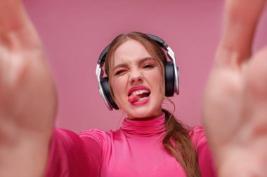 Genç kızıl saçlı kadının portresi. Dilini gösteriyor ve iki eliyle selfie çekiyor. Kulaklık takan komik kız pembe arka planda izole edilmiş bir hediye veriyor. Boşluğu kopyala