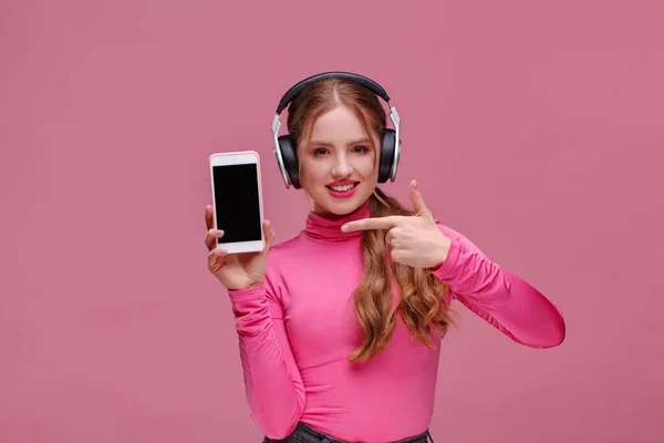 Menina ruiva engraçado mostrando telefone celular tela em branco na câmera. Mulher sorrindo jovem usando fones de ouvido demonstrando exibição de smartphone isolado sobre fundo rosa. Espaço para cópia. App para o seu negócio — Fotografia de Stock