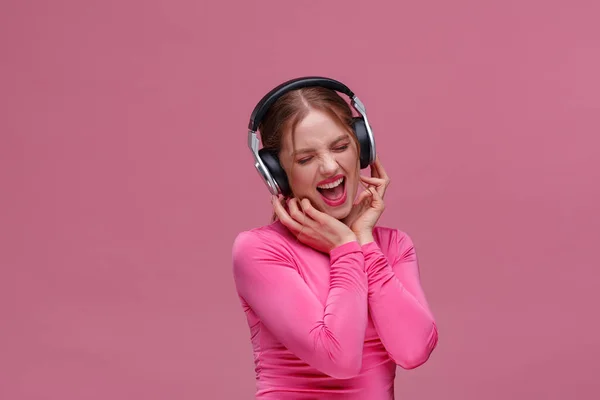 Desfrute de ouvir música. Mulher ruiva bonita em fones de ouvido ouvir música. Menina sorrindo engraçado em fones de ouvido e blusa rosa dançando no fundo rosa. Relaxamento e gestão do stress — Fotografia de Stock