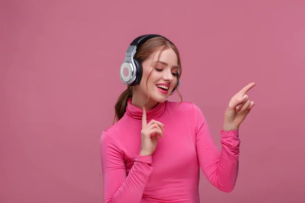 Geniet van muziek luisteren. Mooie jonge roodharige vrouw in koptelefoon die muziek luistert. Grappig lachend meisje in oortjes en roze blouse dansend op roze achtergrond. Ontspanning en stressbeheersing — Stockfoto