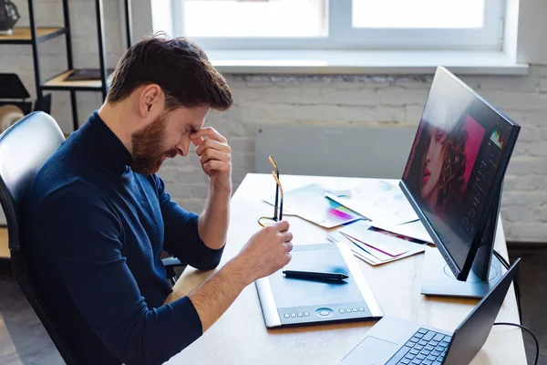 Porträtt av trött grafisk designer som arbetar övertid på kontoret. Stressad arbetare har symptom på ögonpåfrestningar. Retuschera arbetsplatsen i fotostudio. Utbrändhet och överarbete koncept. Kreativ byrå — Stockfoto