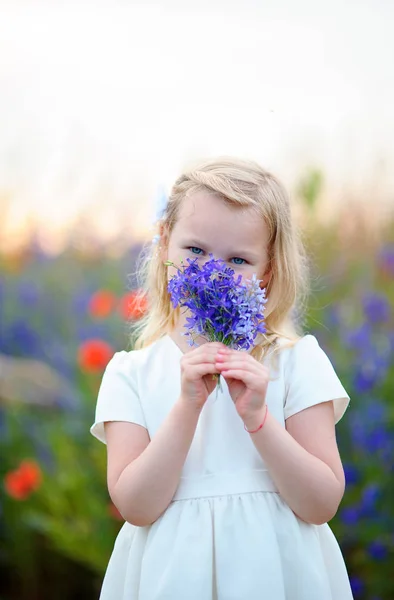 Menina loira feliz vestindo vestido branco cheirando azul selvagem — Fotografia de Stock