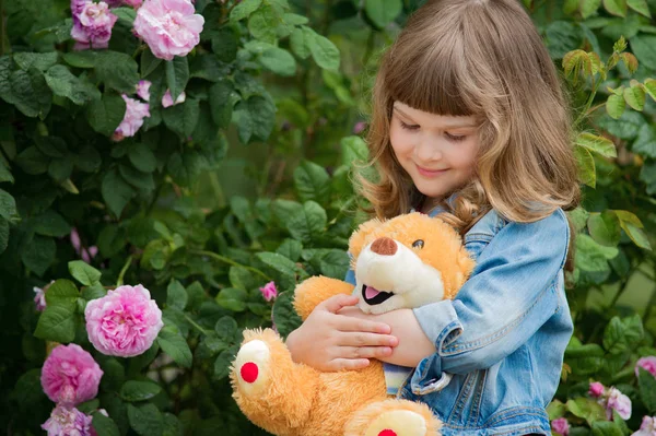 ピンクのバラ公園でテディベアと愛らしい笑みを浮かべて少女. — ストック写真