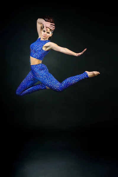 Young woman gymnast in blue suit jumping against a black backgro