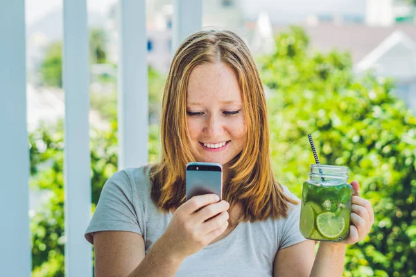 Een jonge vrouw houdt een mason jar in haar hand met een mojito en een smartphone. Lente drinken concept — Stockfoto