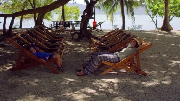 Vrouw en zoon zit in de ligstoelen onder een grote boom — Stockvideo