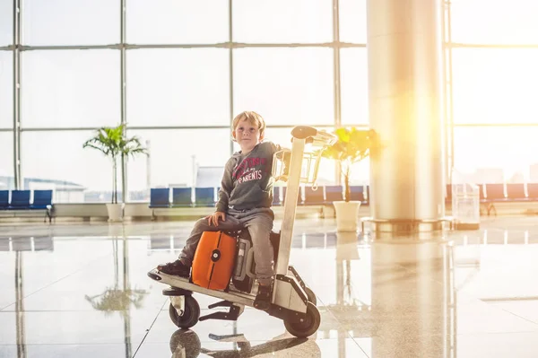 Menino bonito com mala laranja no aeroporto. O rapaz no carrinho e o aeroporto. — Fotografia de Stock