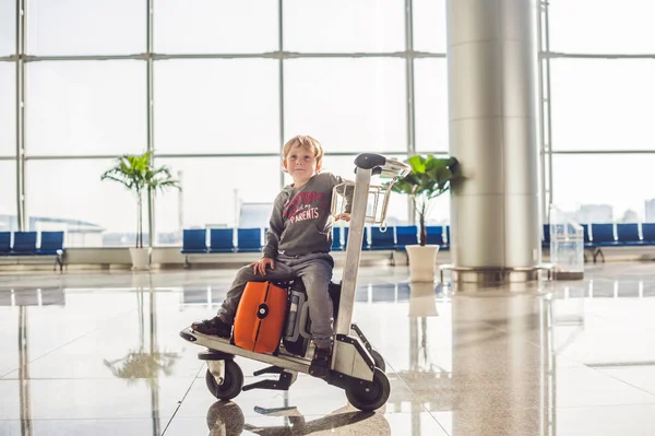 Menino bonito com mala laranja no aeroporto. O rapaz no carrinho e o aeroporto. — Fotografia de Stock