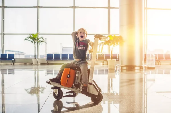 Menino bonito com mala laranja no aeroporto. O rapaz no carrinho e o aeroporto. — Fotografia de Stock