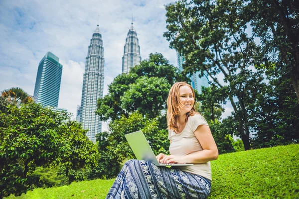 Молодая женщина в повседневной одежде с ноутбуком в тропическом парке на фоне небоскребов. Концепция мобильного офиса — стоковое фото