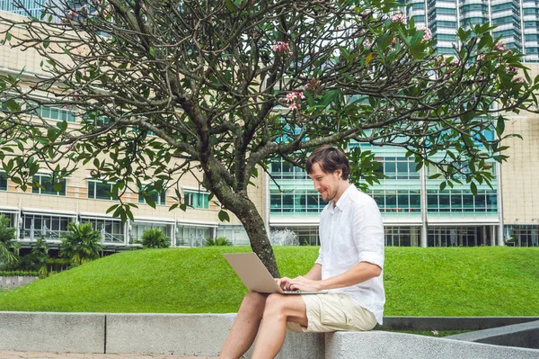 남자 실업가 또는 캐주얼 드레스 마천루의 배경에 열 대 공원에서 노트북을 사용 하 여 학생. 모바일 오피스 개념 — 스톡 사진