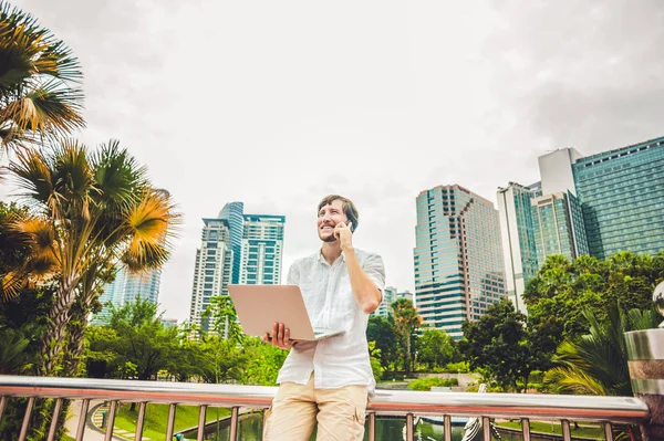 Чоловік-підприємець або студент у повсякденному одязі, використовуючи ноутбук у тропічному парку на тлі хмарочосів. Концепція мобільного офісу — стокове фото