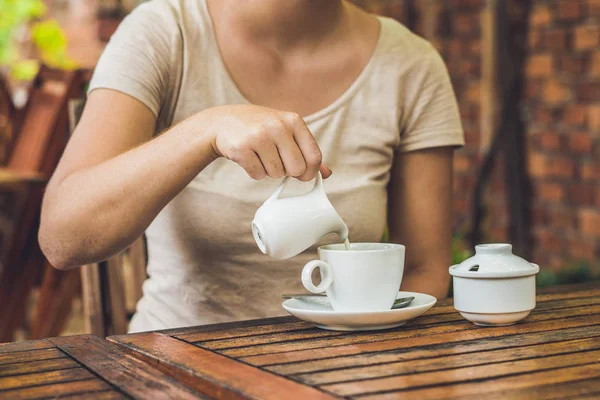 Женская рука наливает молоко в белую чашку кофе в кафе на открытом воздухе — стоковое фото