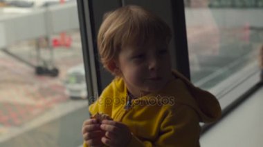 Bir Havaalanı'nda büyük bir pencerenin önünde bir çerez sittting yeme sarı ceketli genç bir çocuk bardak Slowmotion
