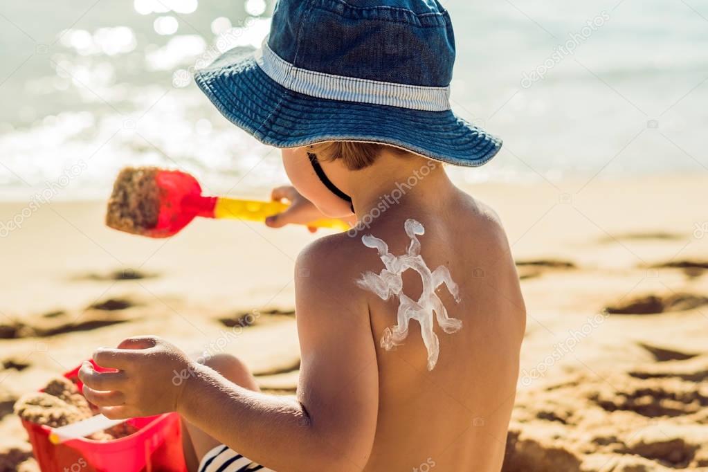 Sun drawing with sunscreen, suntan lotion on baby boy back. Caucasian child is sitting with plastic container of sunscreen and toys on sunny beach. Close up, outdoor.