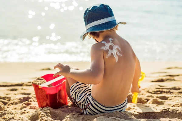 日焼け止め ベビーの男の子の背中に日焼け止めローションを使用した描画の太陽 白人の子は晴れたビーチで日焼け止めとおもちゃのプラスチック コンテナーに座っています 屋外をクローズ アップ — ストック写真