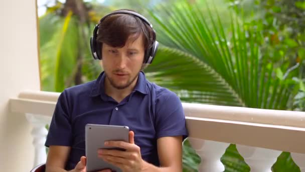 タブレットとテラスで座っている無線のヘッドフォンを使用してビデオ通話でお友達や家族と話して熱帯地方の男 — ストック動画
