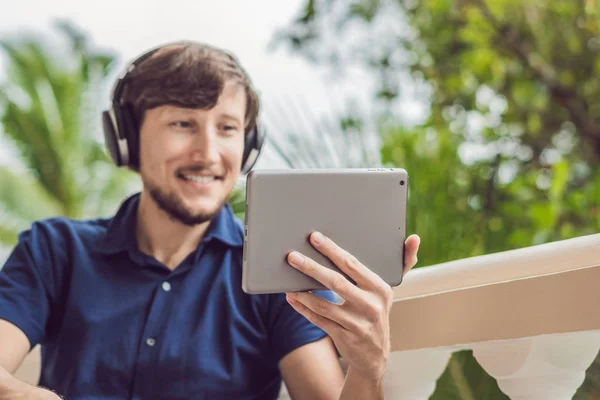 man in the tropics talking with friends and family on video call using a tablet and wireless headphones sitting on the terrace.
