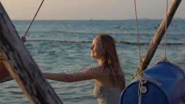 Jovem mulher balança seu filho feliz em um baloiço de madeira em um mar em um horário de pôr do sol — Vídeo de Stock