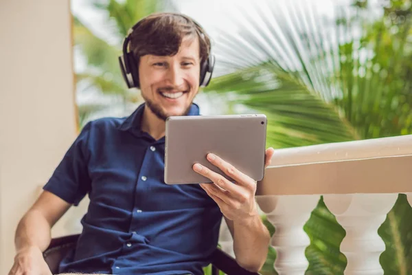 man in the tropics talking with friends and family on video call using a tablet and wireless headphones sitting on the terrace.